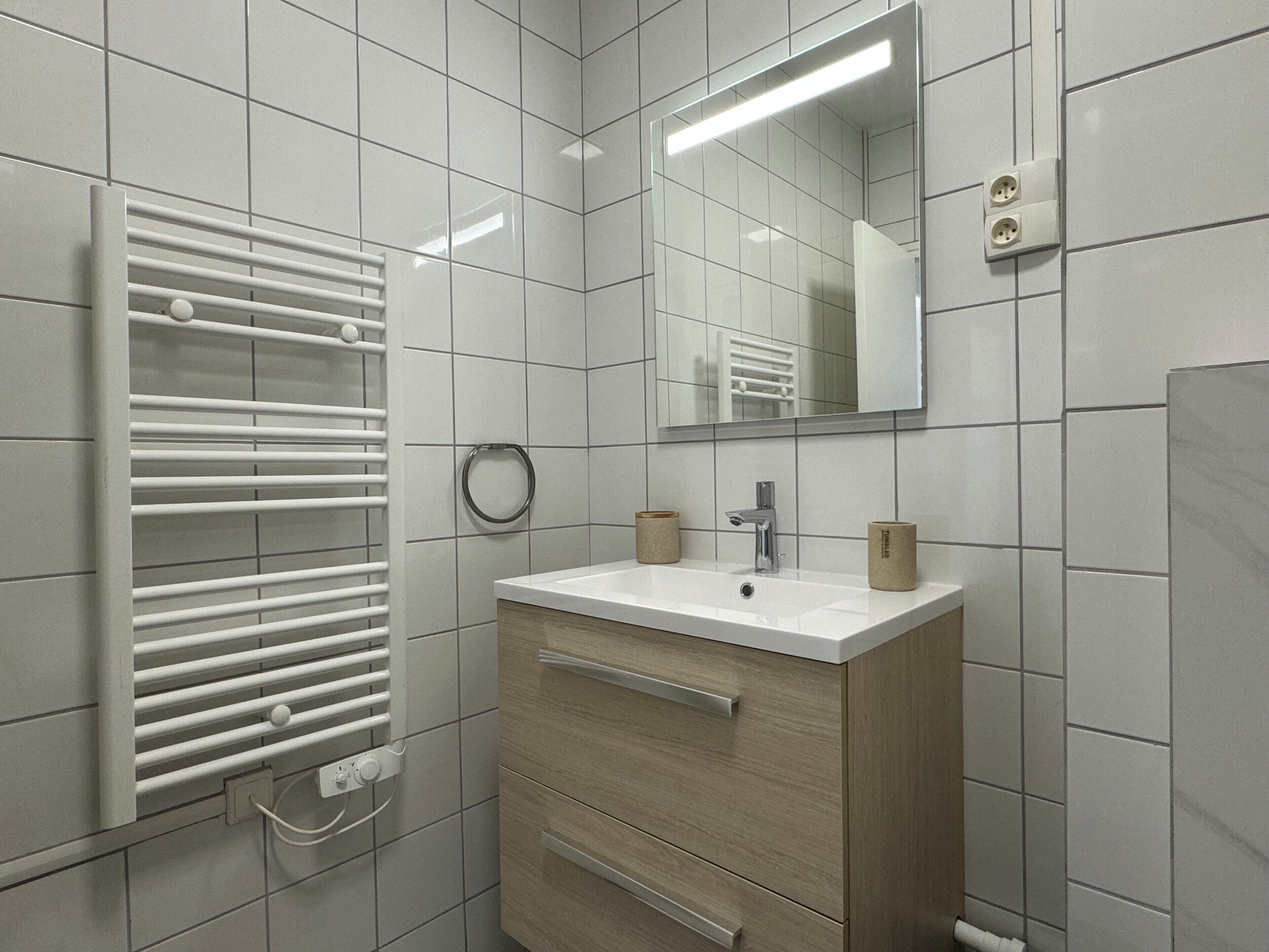 Une salle de bain moderne avec un mur recouvert de carreaux blancs, un lavabo blanc avec un meuble-vasque en bois, un porte-serviettes et un miroir.