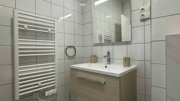 Une salle de bain moderne avec un mur recouvert de carreaux blancs, un lavabo blanc avec un meuble-vasque en bois, un porte-serviettes et un miroir.