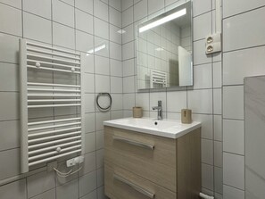Ein modernes Badezimmer mit weiß gefliesten Wänden, einem weißen Waschbecken mit Holzschrank, einem Handtuchhalter und einem Spiegel.