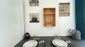 A dining area with a table set for two, featuring a modern design with geometric patterns on the plates. There are two wine glasses, a water glass, and a kettle on the table. The wall behind the table has a wooden shelf with bottles and a framed quote about cooking. - Les Capucines - Tournon-sur-Rhône (Tournon-sur-Rhône)