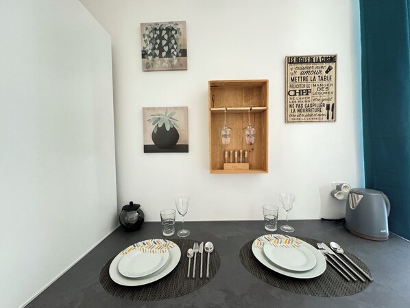 A dining area with a table set for two, featuring a modern design with geometric patterns on the plates. There are two wine glasses, a water glass, and a kettle on the table. The wall behind the table has a wooden shelf with bottles and a framed quote about cooking.