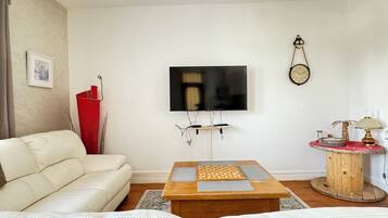 Un salon comprenant un canapé, une table basse avec un jeu d’échecs, un téléviseur fixé au mur, une horloge et une lampe sur une table d’appoint.