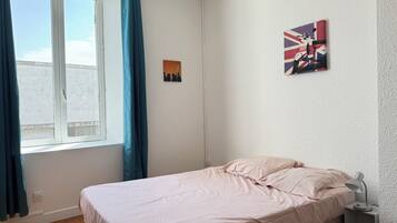 Une chambre à coucher avec un lit, une fenêtre avec des rideaux, un tableau accroché au mur et une table de chevet avec une lampe.