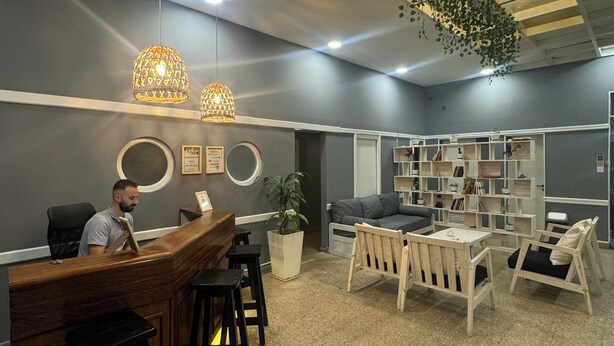 Un área de recepción con un escritorio de madera, taburetes negros, una zona de estar con sofá y sillones, una planta en maceta, y un techo con vigas a la vista y luces colgantes.