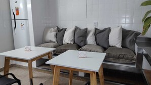 Una kitchenette con un refrigerador blanco, una mesa con base de madera y un sofá gris con varios cojines.