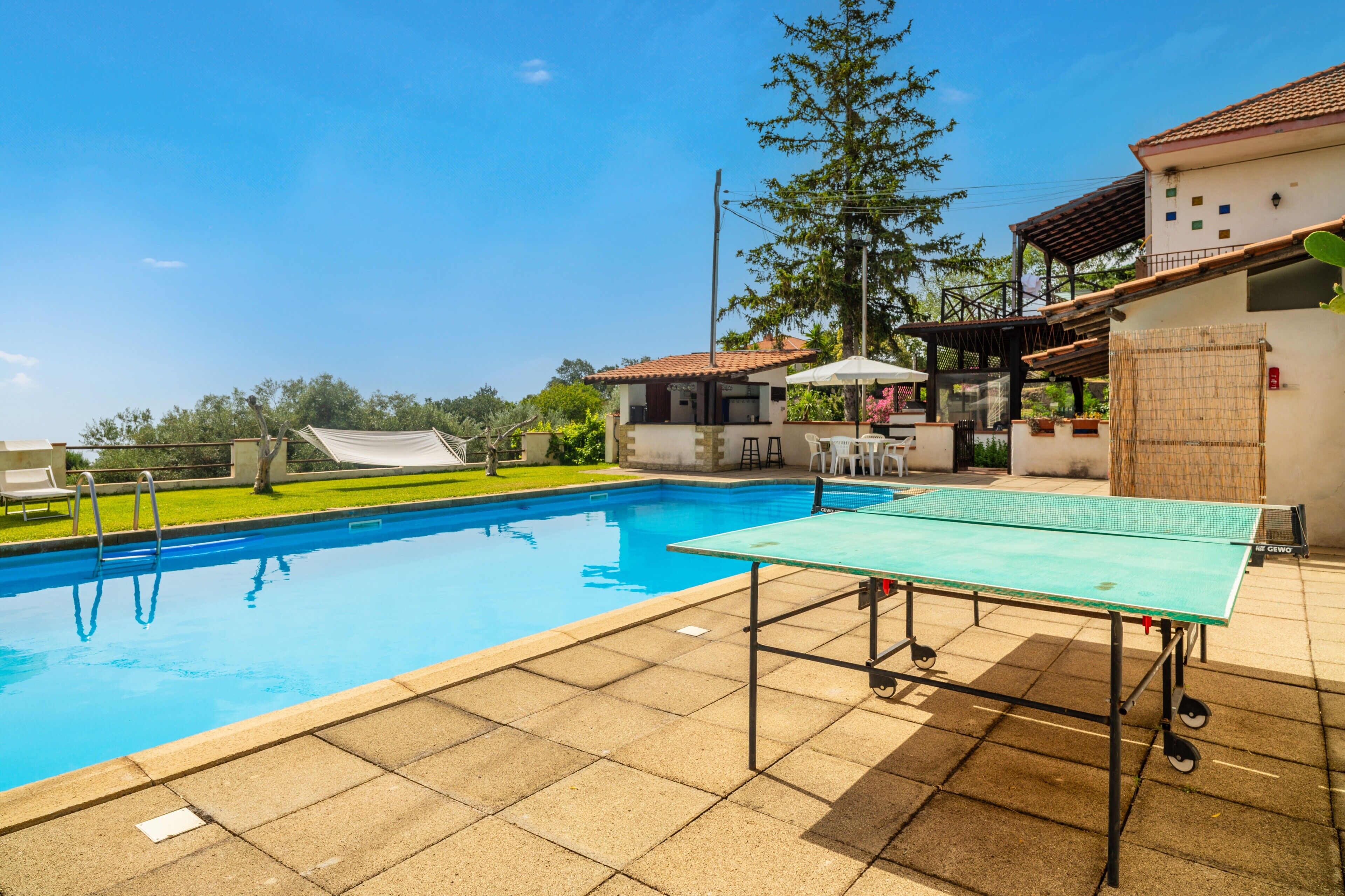 Una piscina con tavolo da ping pong, un'area salotto coperta e un edificio con tetto in tegole.