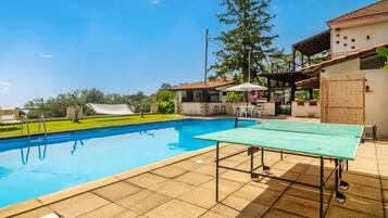 Una piscina con tavolo da ping pong, un'area salotto coperta e un edificio con tetto in tegole.