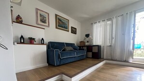 Ein Wohnzimmer mit einem blauen Sofa, Holzboden, einer weißen Wand mit gerahmten Bildern und einem Fenster mit weißen Vorhängen.