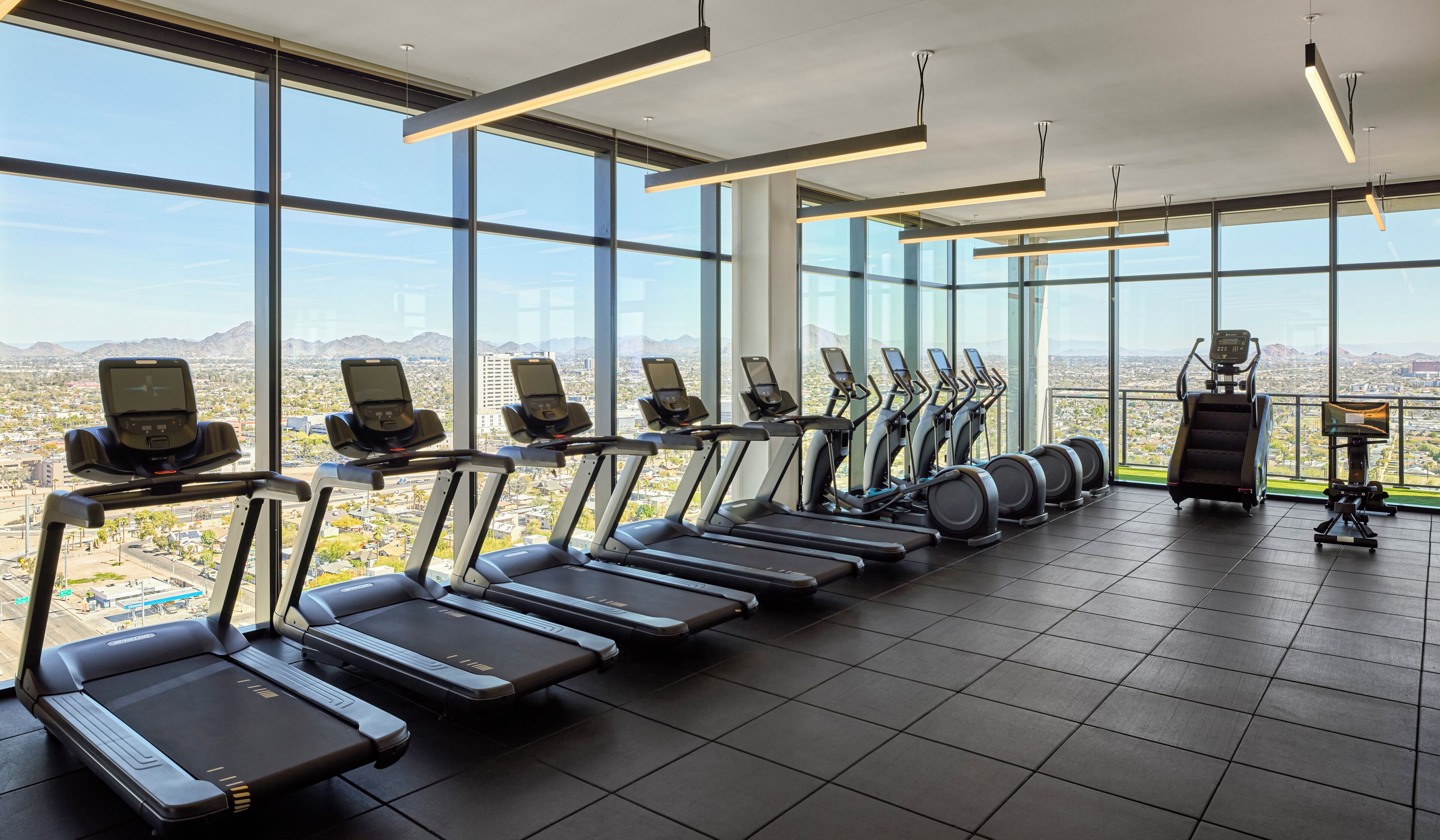 Un gimnasio con cintas de correr y elípticas, amplias ventanas con vistas a la ciudad e iluminación moderna.