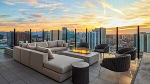 Eine Dachterrasse mit Sofagarnitur, Stühlen und einer Feuerstelle mit Blick auf eine Stadtlandschaft bei Sonnenuntergang.