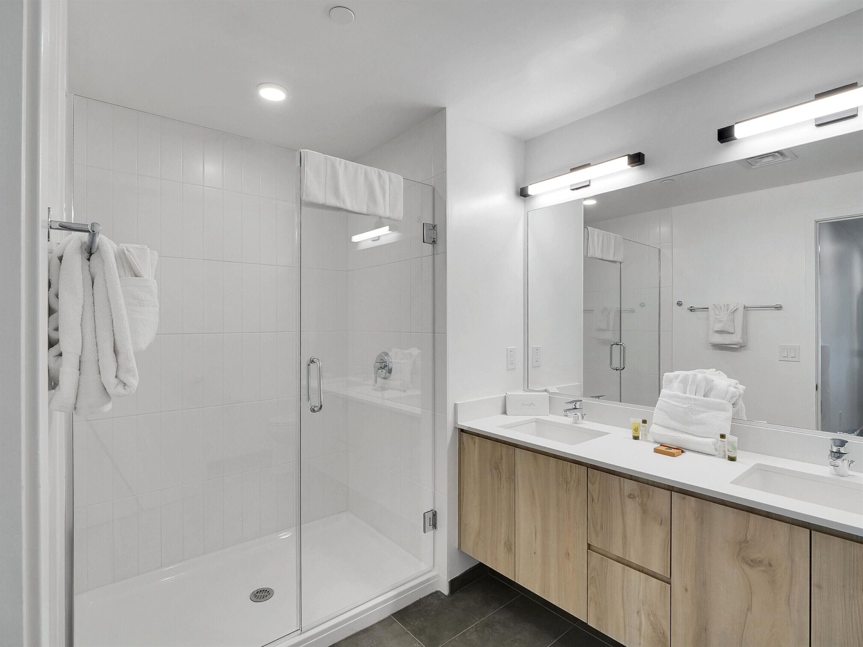 Un baño moderno con ducha envuelta en vidrio, un espejo grande y un tocador de madera con lavabo.