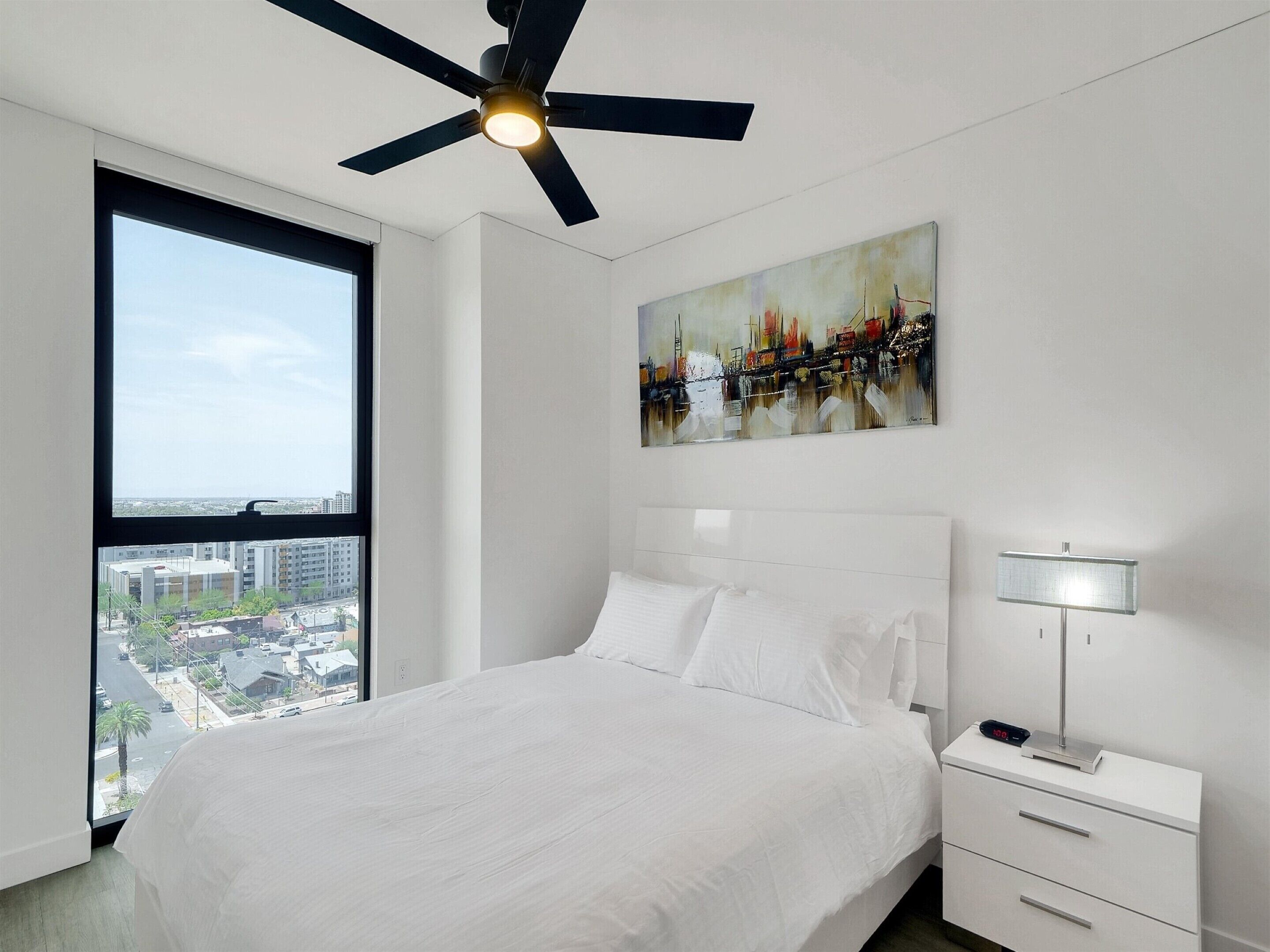 A modern bedroom with a large bed, a ceiling fan, a nightstand with a lamp, and a painting on the wall.