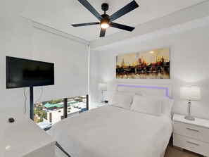 A modern hotel room with a bed, a flat-screen TV, a ceiling fan, a cityscape painting, and a view of the cityscape through large windows.