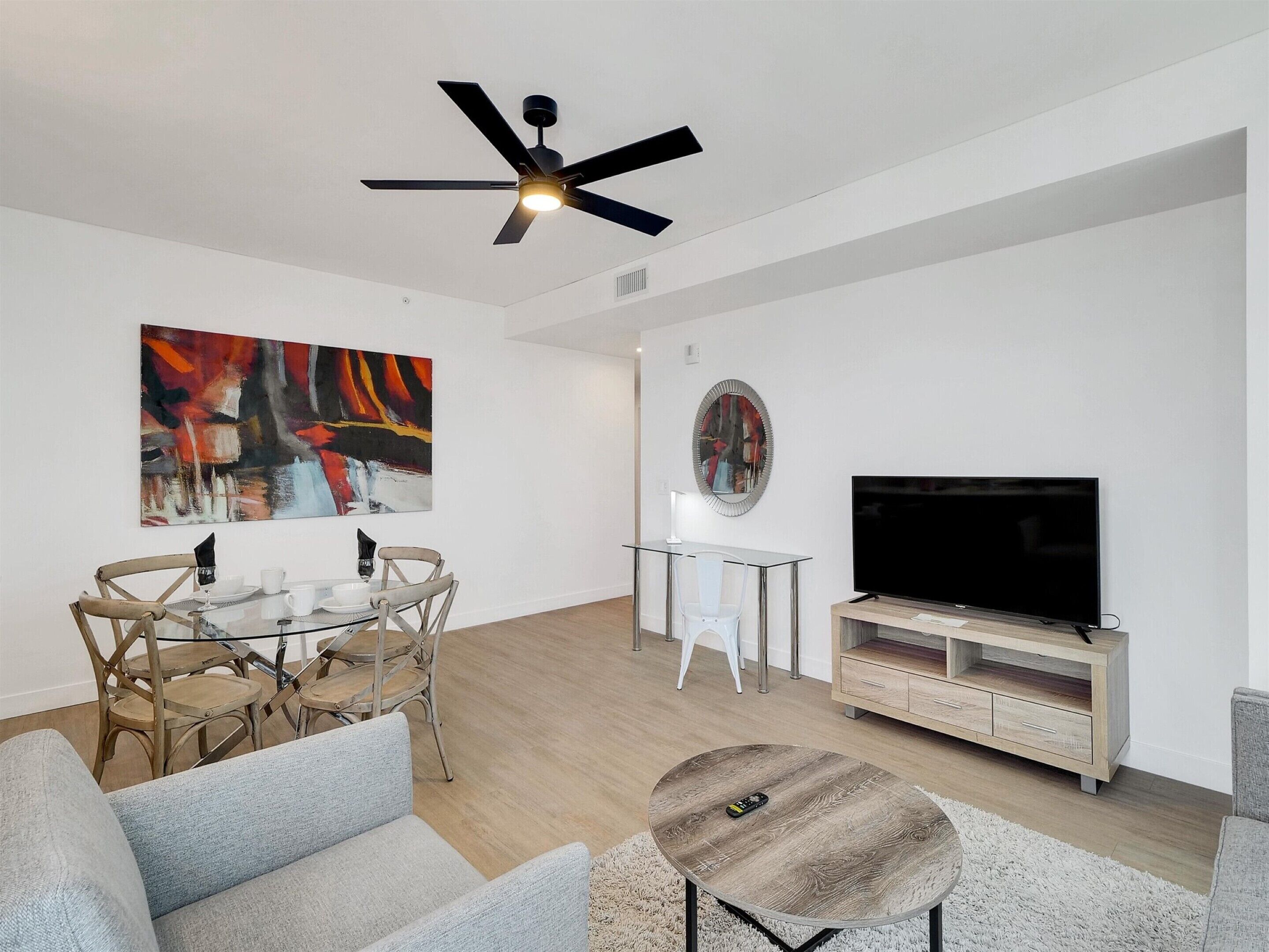 Een moderne woonkamer met een flatscreen televisie, een plafondventilator, een eethoek met een glazen tafel en stoelen, en een groot abstract schilderij aan de muur.