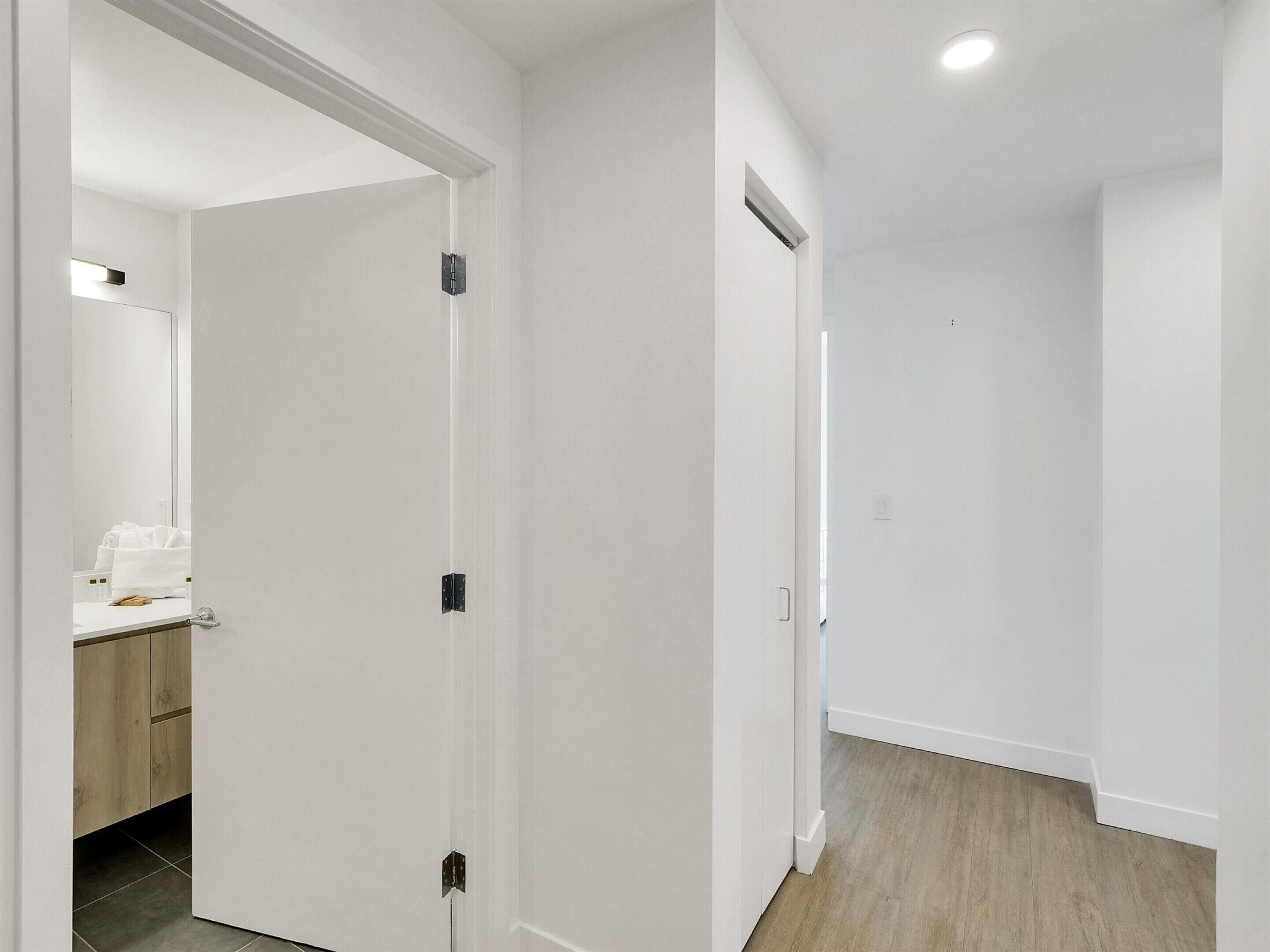 A clean, white-walled hallway with a wooden floor, a bathroom with a mirror and towel, and a door with a handle.
