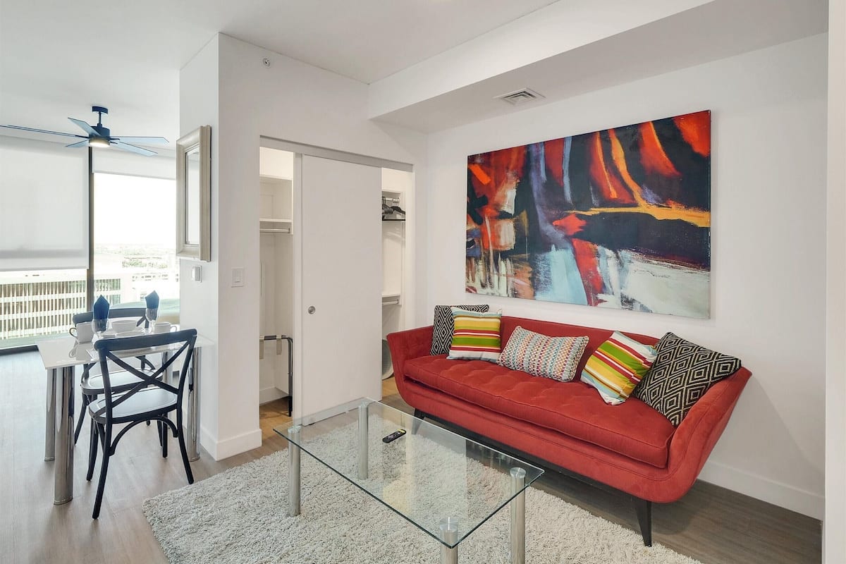 A modern living room with a glass coffee table, a red sofa with patterned cushions, and a large abstract painting on the wall.