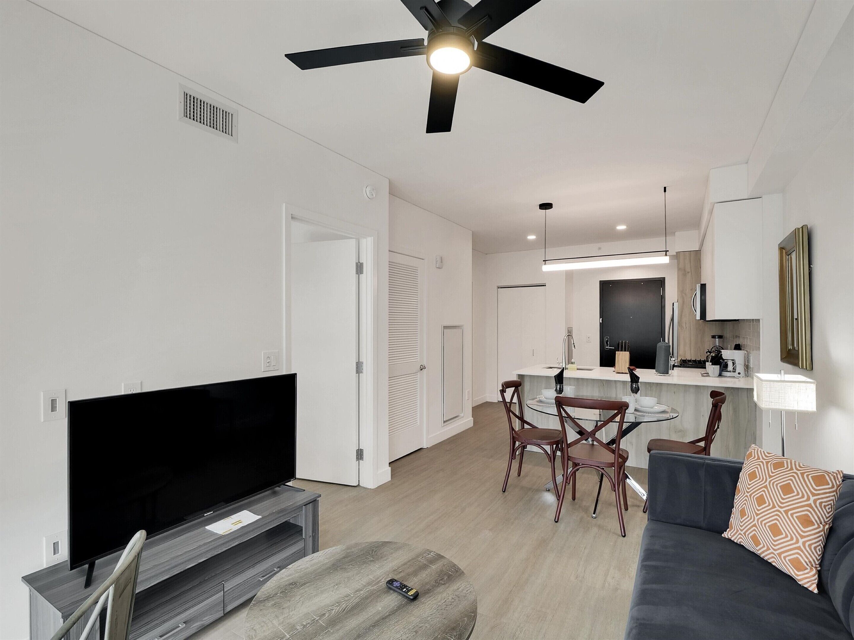 A modern living room with a flat-screen TV, a ceiling fan, a dining area with a table and chairs, and a kitchen area with a sink and cabinets.