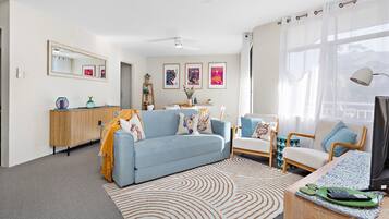 A modern living room with a sofa, armchairs, a coffee table, and a TV. There is a dining area with a table and chairs, and a bookshelf. The room has large windows with sheer curtains.