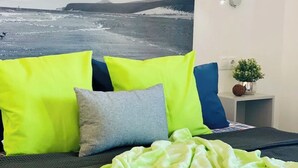 Un lit recouvert d’une couverture verte, deux oreillers aux couleurs vives et une décoration murale représentant un paysage marin.