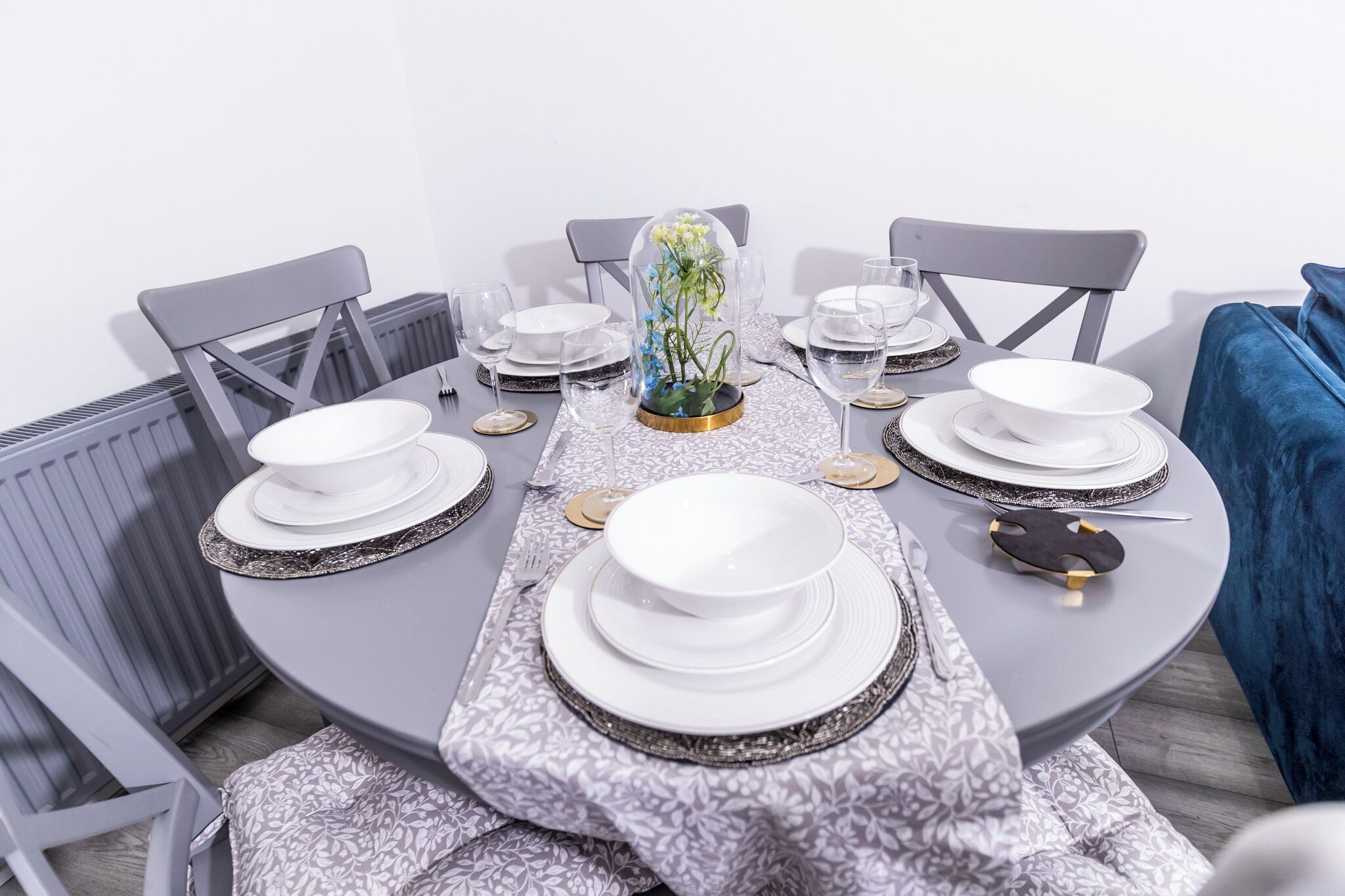 Uma mesa de jantar posta com pratos, tigelas e talheres brancos, rodeada por cadeiras cinzentas.