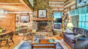 Un acogedor interior de cabaña de troncos con chimenea de piedra, mobiliario de madera y un televisor.