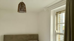 Una camera da letto con un letto grande, due cuscini decorativi, una lampada a sospensione, una finestra con tende e una piccola pianta in vaso.
