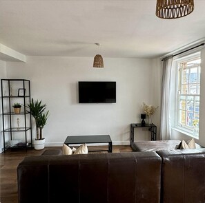 A living room with a black leather sofa, a TV mounted on the wall, a black table, a wicker pendant light, and a window with curtains.