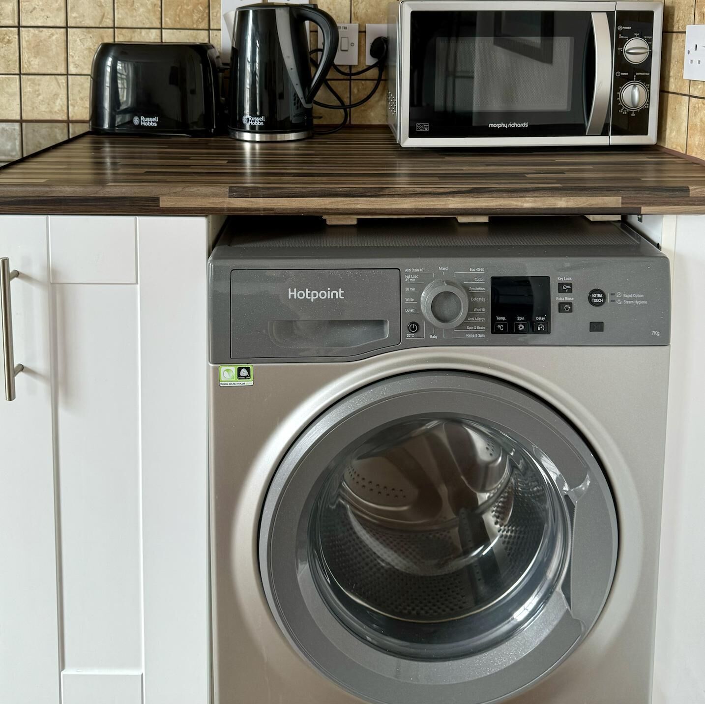 Un appareil compact combinant lave-linge et sèche-linge, doté d’un panneau de commande, ainsi qu’une bouilloire et un four à micro-ondes placés sur le plan de travail.
