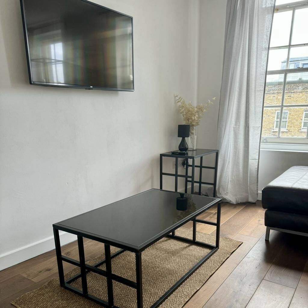 Een moderne woonkamer met een flatscreen televisie aan de muur, een zwarte salontafel, een klein bijzettafeltje met een lamp, en uitzicht op een gebouw door een raam met witte gordijnen.