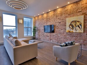 Ein modernes Wohnzimmer mit Backsteinwand, Flachbildfernseher, Sofa, Sessel und großem Fenster.