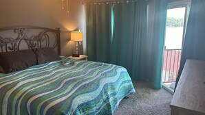 Habitación de hotel con una cama, una mesita de noche con lámpara, ventilador de techo y vistas al exterior a través de una ventana.
