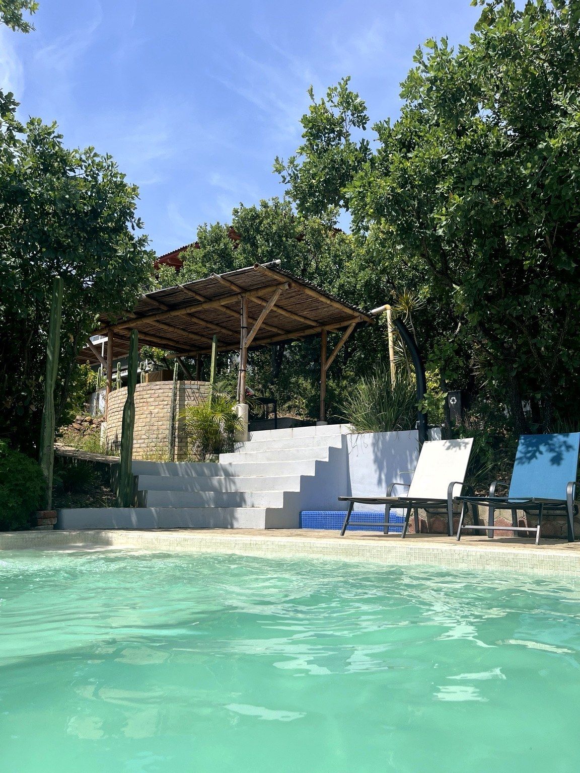 Ett poolområde med en träpergola, trappsteg som leder till en överbyggd sittplats och solstolar.