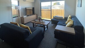 A living room with a dark-colored sofa, a coffee table, and a TV. There is a sliding glass door leading to a backyard. - Home away from home in Brighton (Brighton)