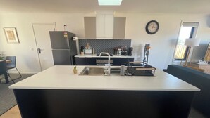 A modern kitchen with a central island, a refrigerator, a clock, and a window with blinds. - Home away from home in Brighton (Brighton)