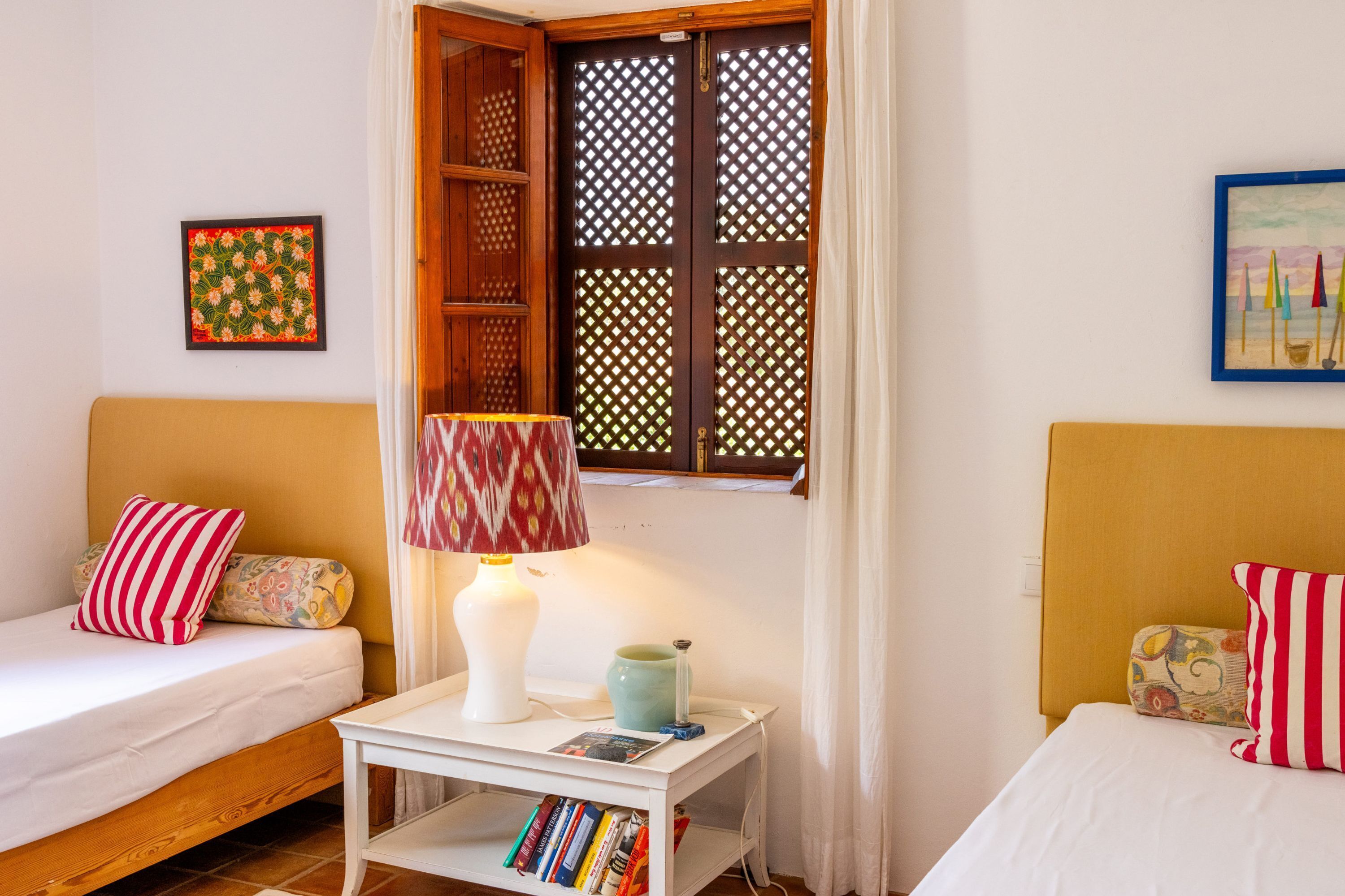 A room with two single beds, a small table with a lamp, a bookshelf, and a window with a lattice screen.