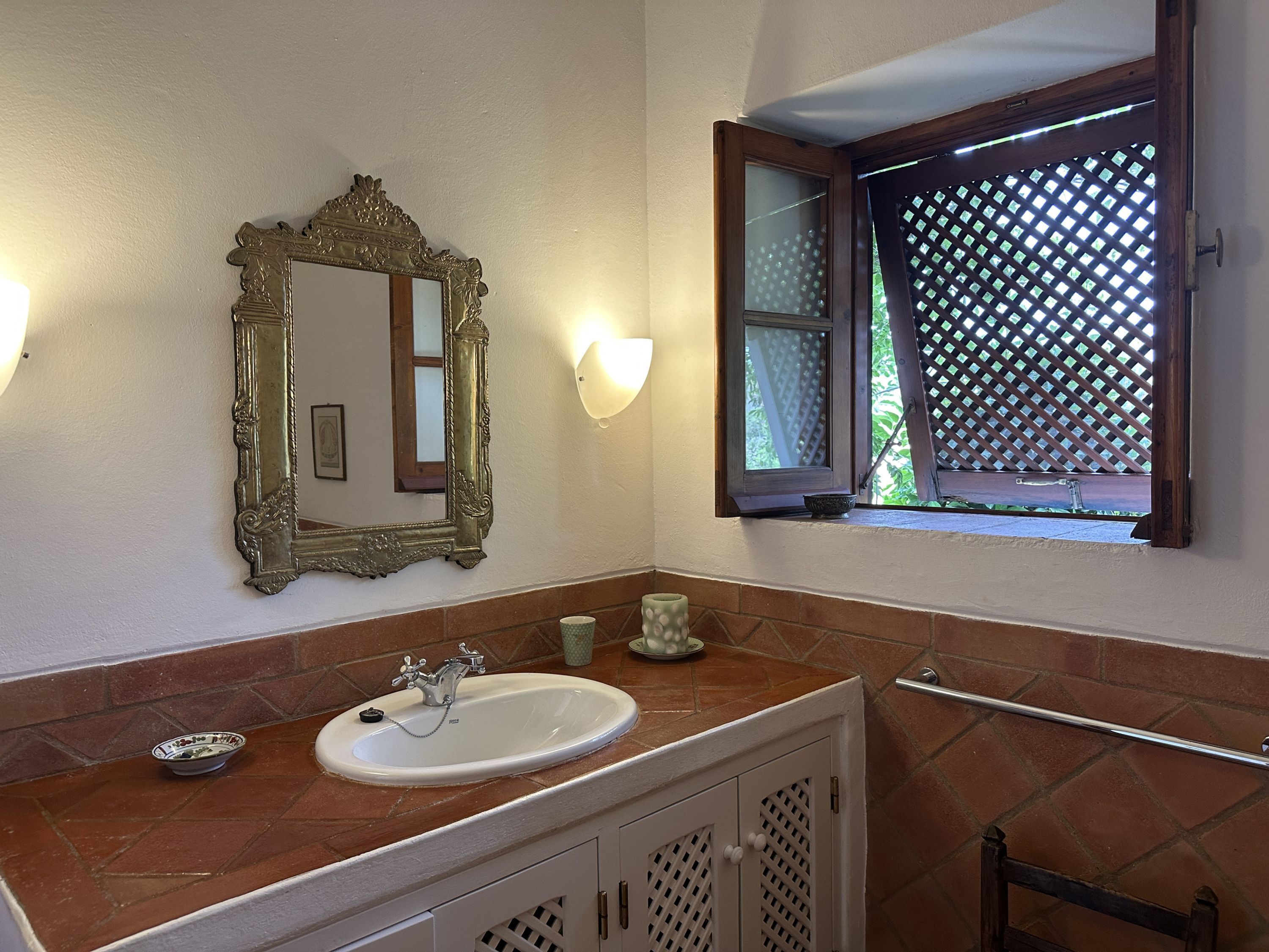 Une salle de bain avec un lavabo, un miroir et une fenêtre ornée d’un treillis.