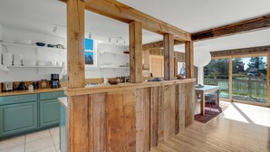 Une cuisine avec des poutres en bois, un coin repas avec une table et des chaises, et une vue sur l’extérieur.