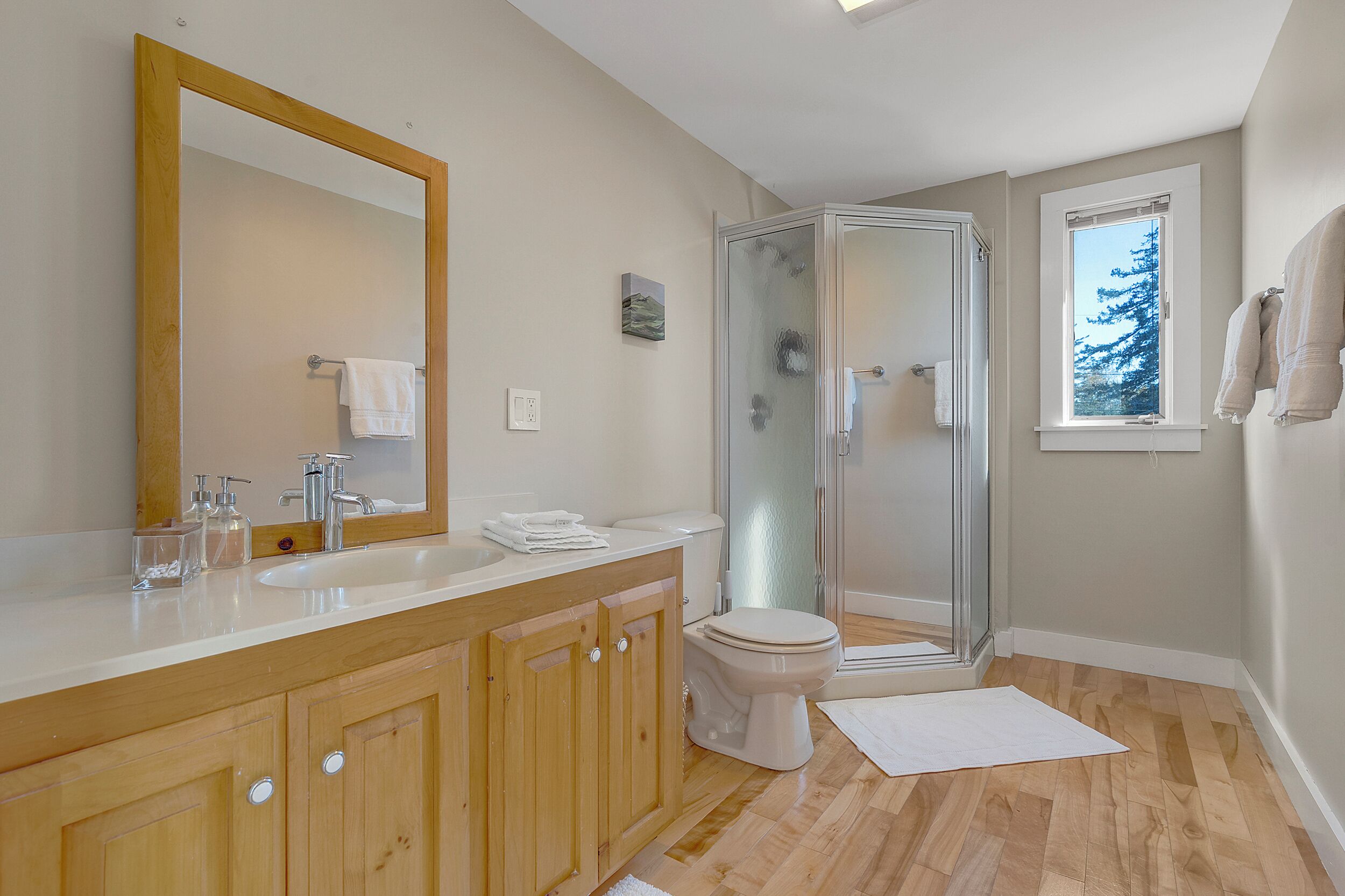 A bathroom with a wooden vanity, a large mirror, a toilet, a glass shower enclosure, and a window with a view of trees.