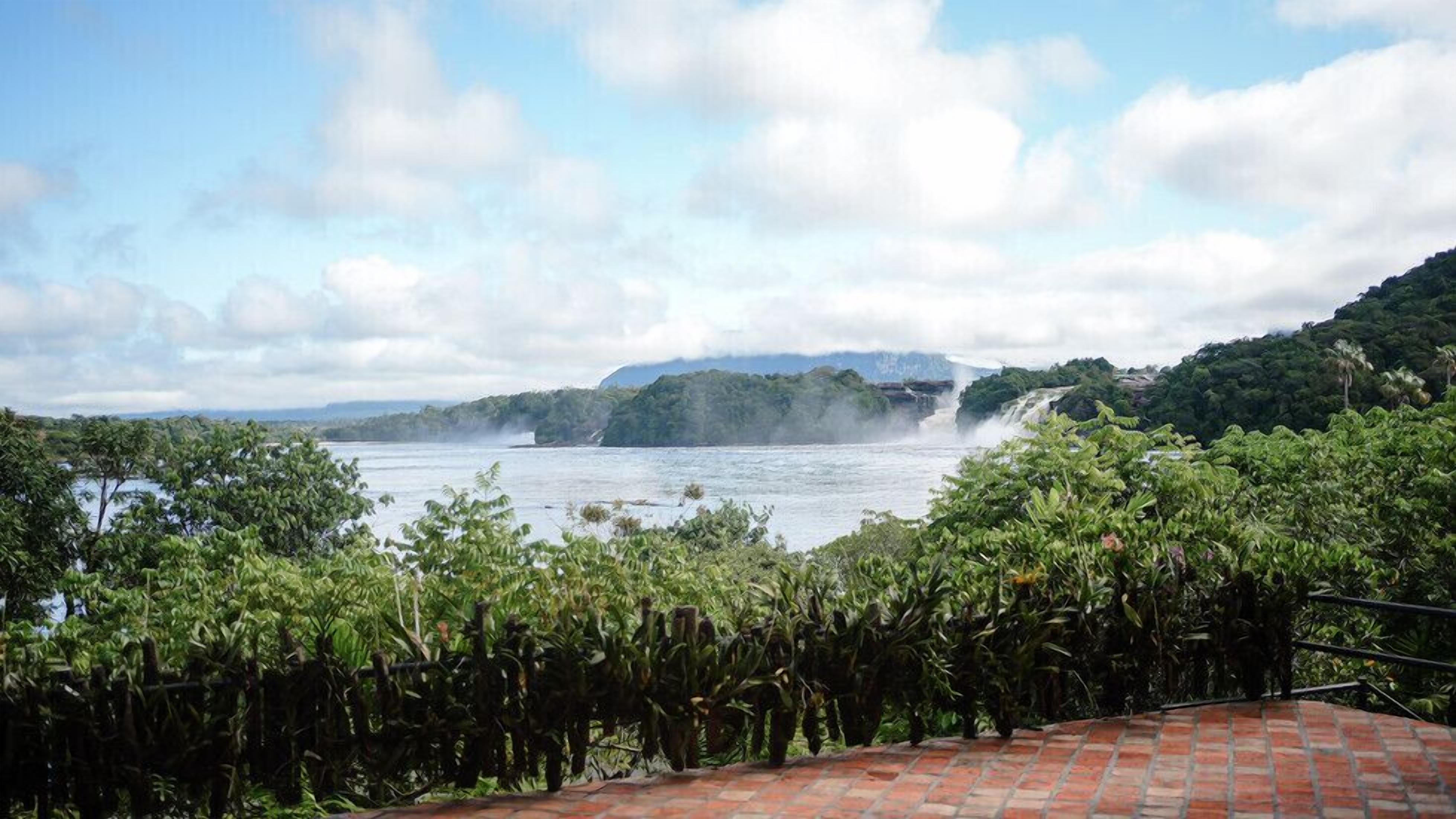 A view of a river with waterfalls, surrounded by lush greenery and a mountainous backdrop.