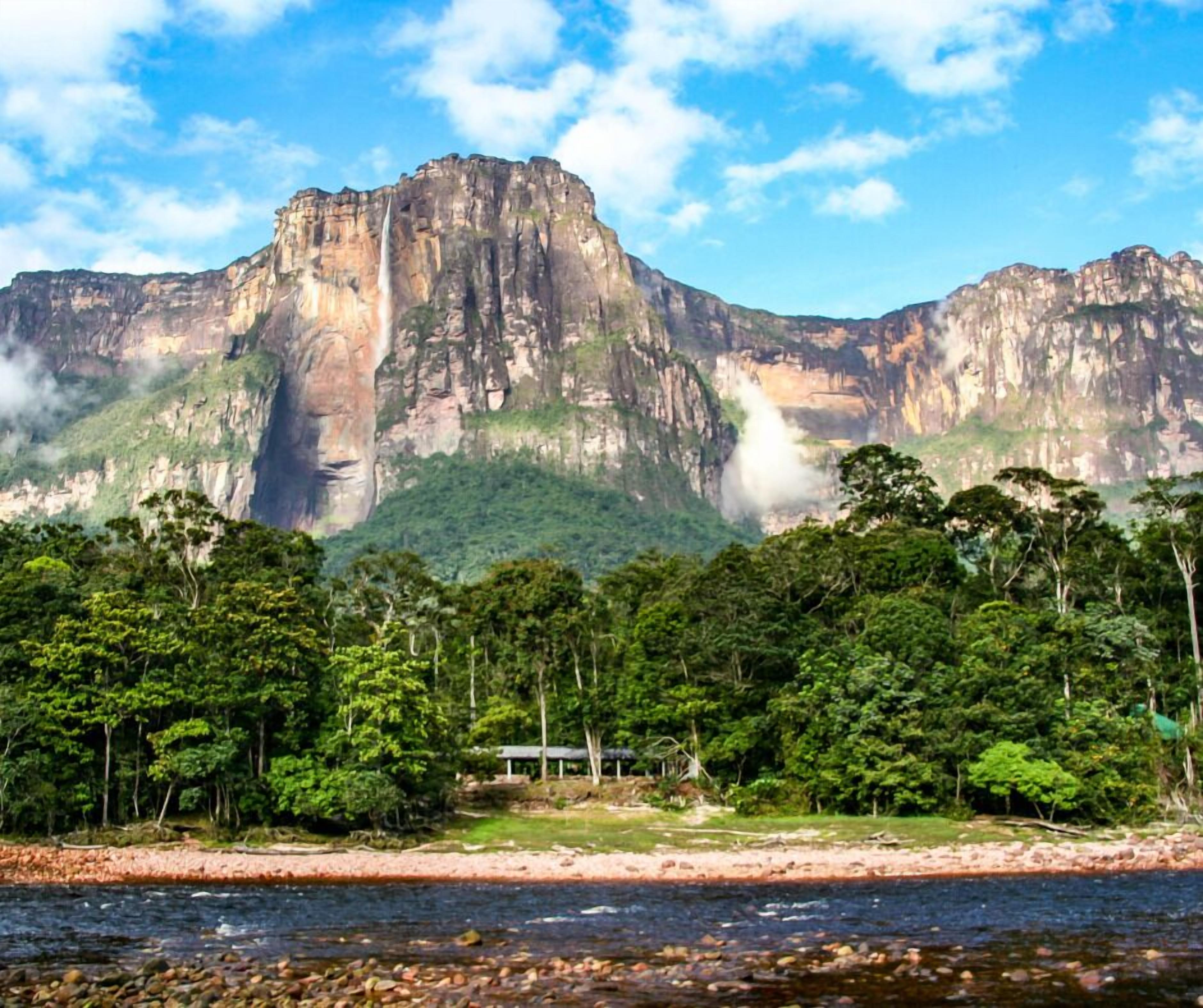 A river with a rocky shoreline, a forested area with a small structure, and a mountain with a waterfall in the background.