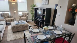 A dining area with a glass table set for four, surrounded by patterned armchairs and a sofa. A flat-screen TV is mounted on the wall above the seating area.