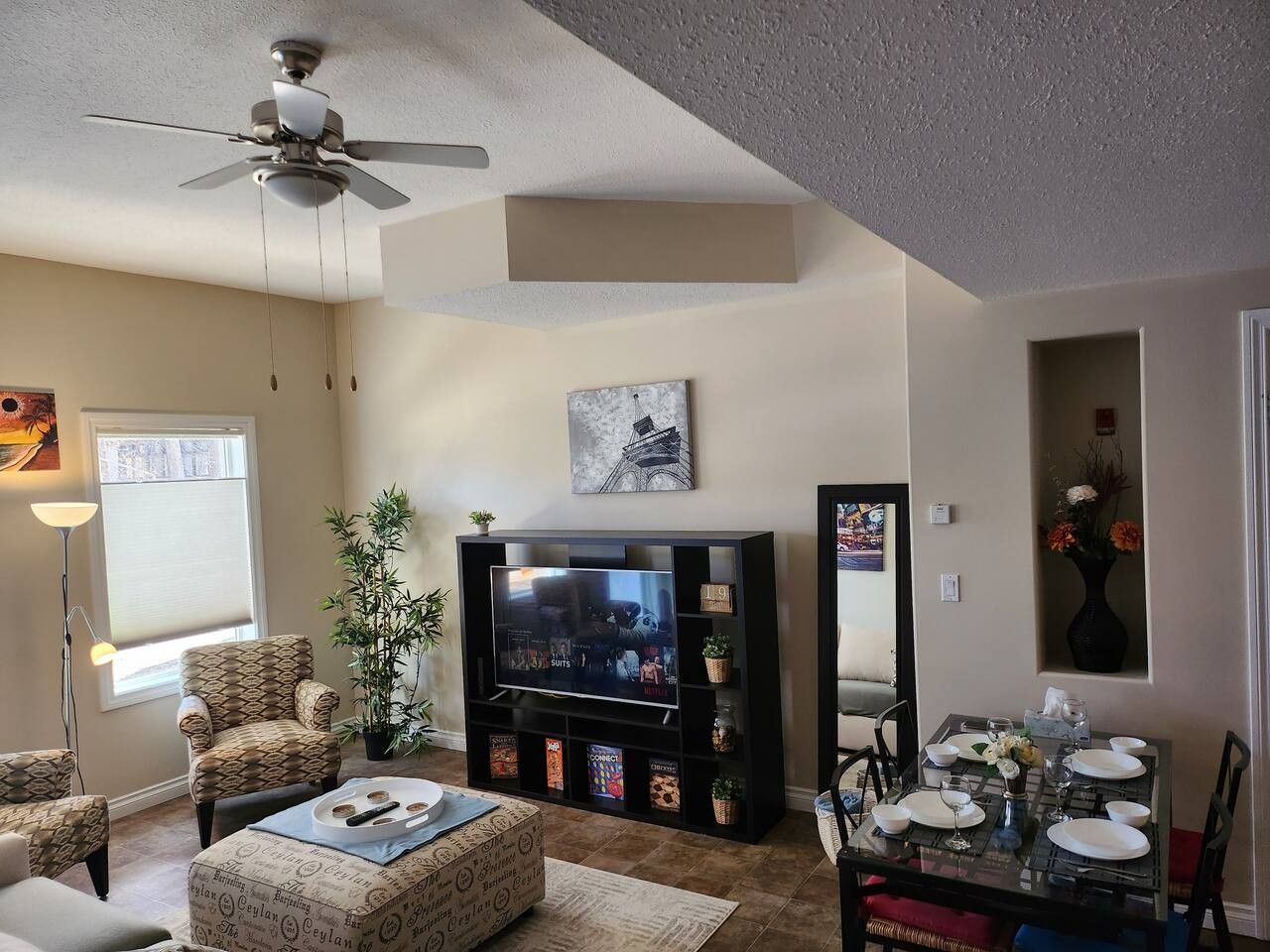 A living room with a flat-screen TV, a ceiling fan, a dining table set for four, and a bookshelf.