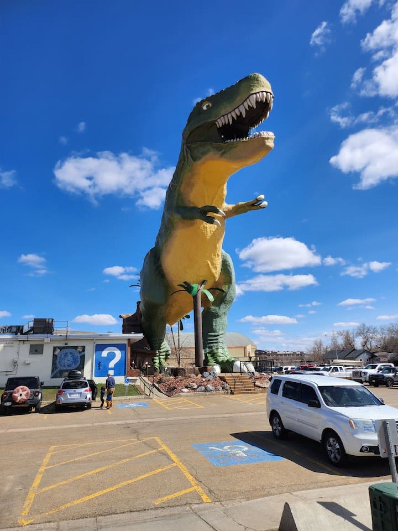 Een grote opblaasbare dinosaurus in een parkeerplaats, met auto's en een gebouw op de achtergrond.
