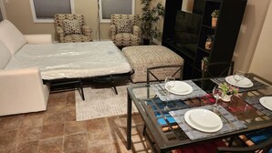 A living room with a sofa, ottoman, patterned chairs, a glass-top dining table set with plates and glasses, a television, and a bookshelf.