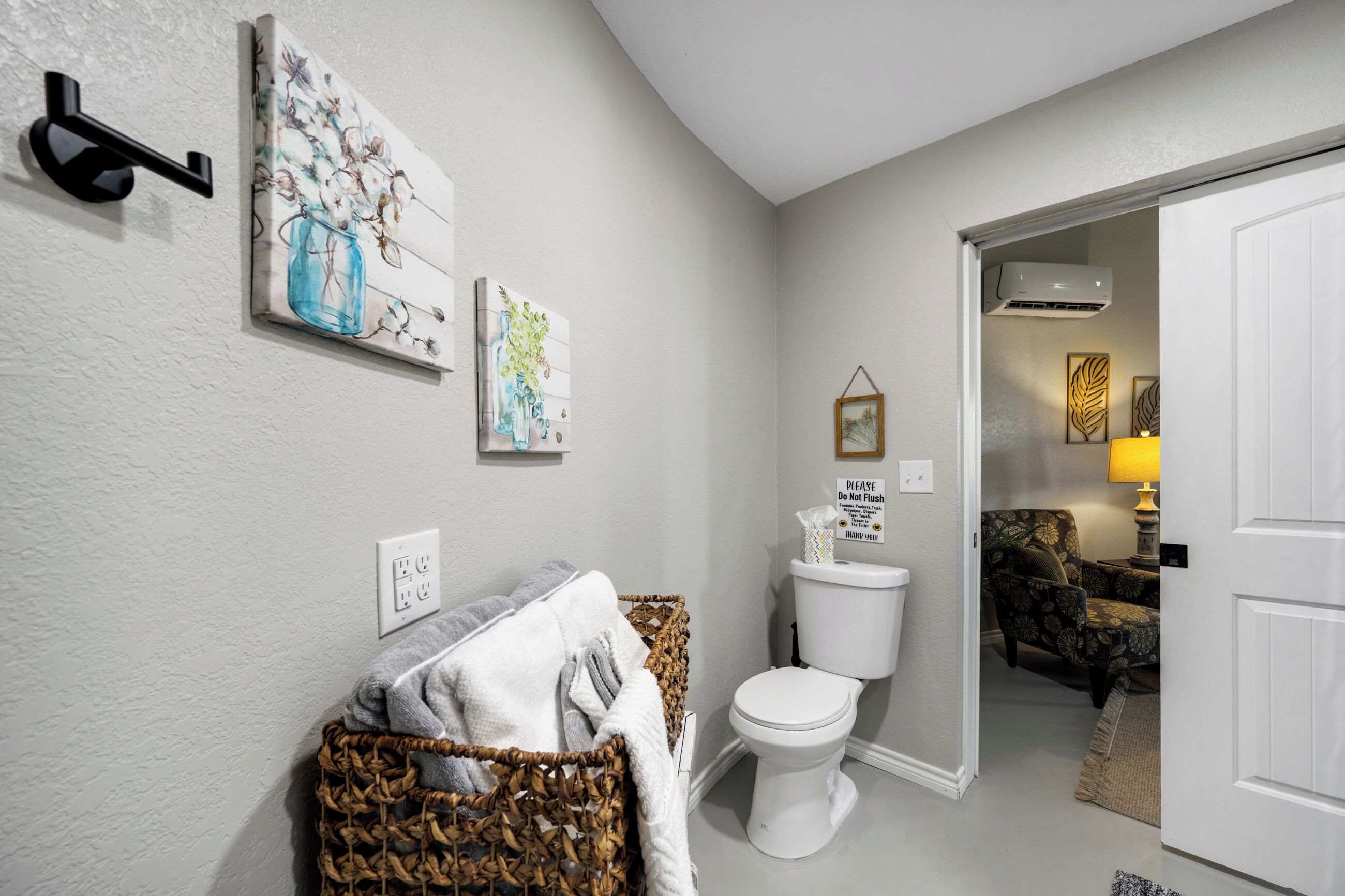A bathroom with a toilet, a basket of towels, and wall-mounted artwork.