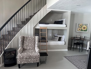 Ett kompakt bostadsutrymme med en trappa, våningssängar, en mönstrad fåtölj, ett litet bord och en matta.