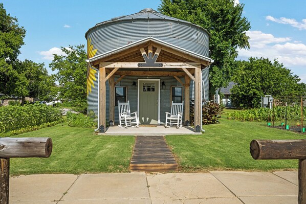 A cylindrical building with a porch and two rocking chairs, surrounded by a well-maintained lawn and trees. - The Painted Silos - The Sunflower Bin. Elgin, Ft Sill and Lawton areas. (Elgin)