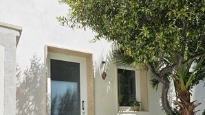 Ein Haus mit weißer Fassade, einer Glastür und einem kleinen Fenster mit Pflanze. Vor der Tür stehen zwei Blumentöpfe.