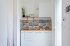 A compact kitchen with white cabinets, a wooden countertop, a backsplash with leaf-patterned tiles, a small shelf with a plant, and a white wall-mounted microwave. - Holiday Home 'Levante' with Shared Terrace, Wi-Fi and Air Conditioning (San Cesario di Lecce)