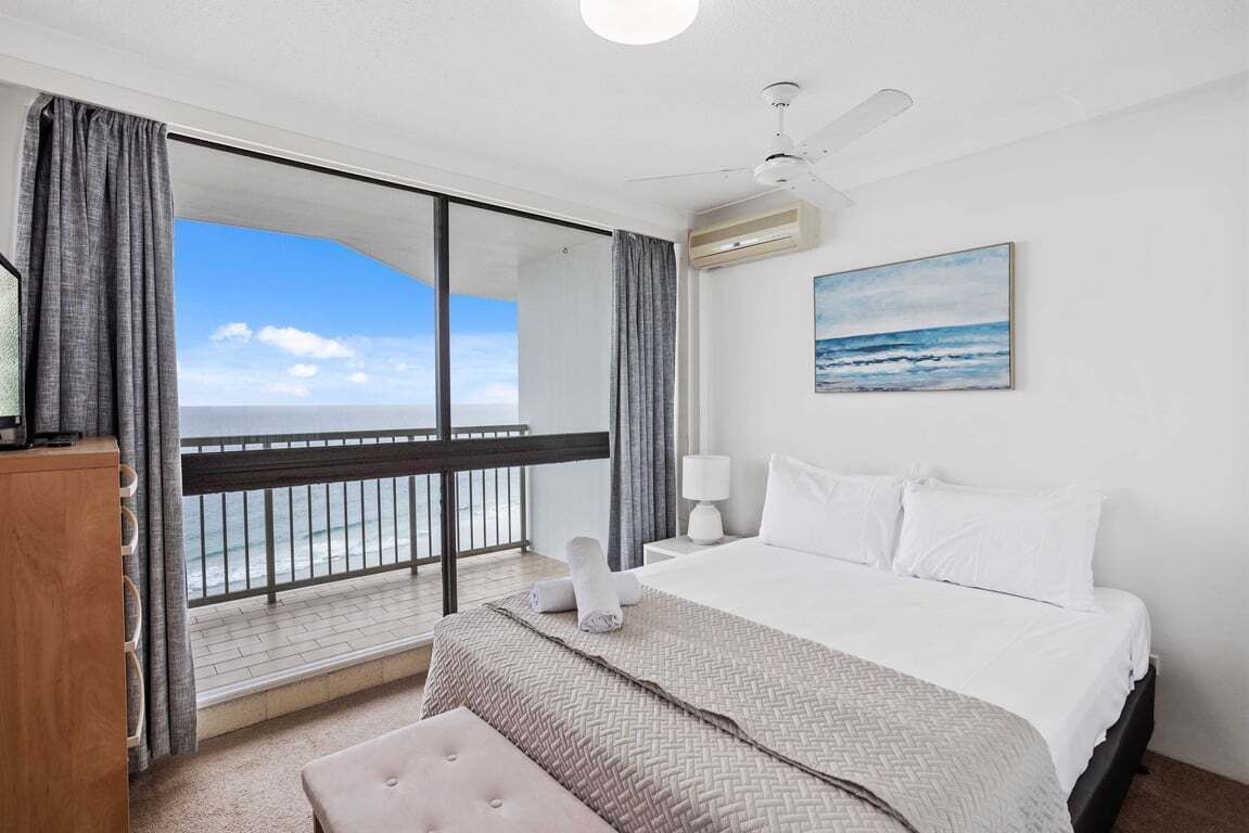 A hotel room with a large bed, a ceiling fan, a painting of the ocean, and a view of the sea through a balcony.